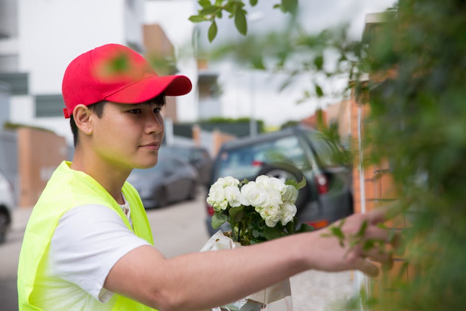 A young male florist wearing a red cap and a high-visibility yellow vest is placing a bouquet of fresh white flowers, likely hydrangeas or roses, against a garden wall. The arrangement showcases vibra