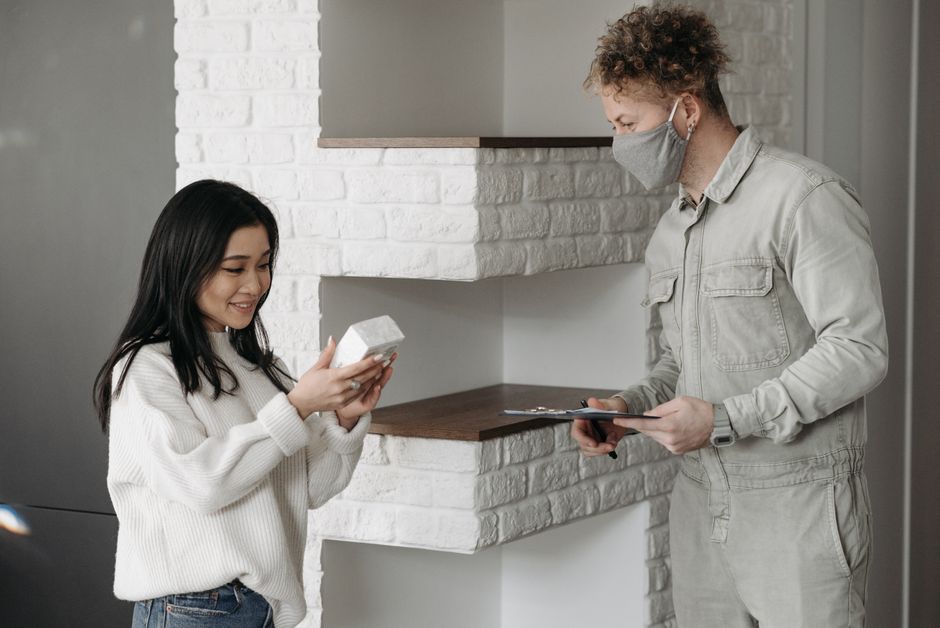 A woman with long dark hair, smiling and holding a small wrapped floral gift, receives a flower delivery from a delivery person wearing a grey face mask and beige jumpsuit. The delivery person is stan