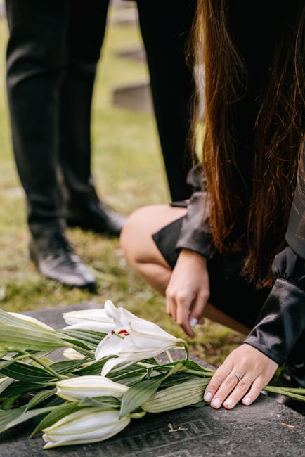 A woman with long brown hair, dressed in dark clothing, kneels on the ground arranging a bouquet of white lilies with green leaves, which are laid on a flat surface in a memorial or funeral setting. T