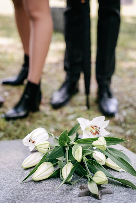 A bouquet of white lilies with large, open blooms and elongated leaves, resting on a stone surface. The bouquet includes closed buds and is partially wrapped in clear cellophane, with some green folia
