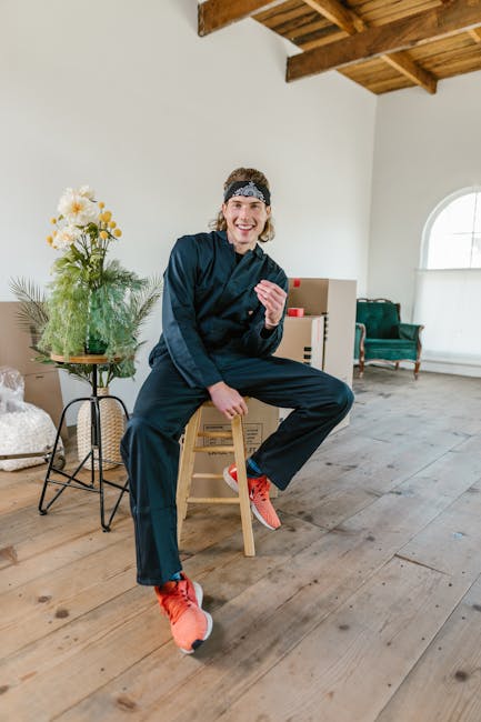 A young woman with light skin and a cheerful expression, wearing a navy-blue tracksuit, bright orange running shoes, and a patterned headband, is sitting on a small wooden stool inside a bright room w