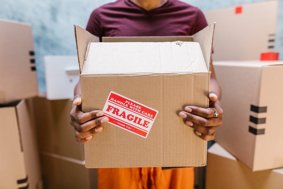 A person holding an open cardboard box marked with a red and white 'Fragile' handling sticker, with their hands visible and wearing a ring on their left ring finger. The individual is standing in a st